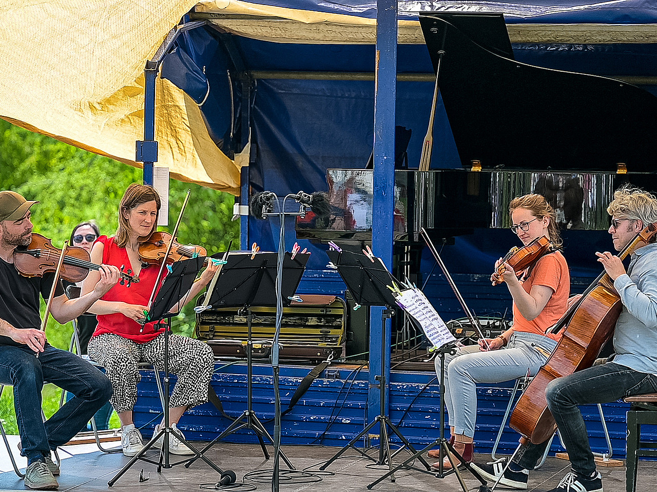Ein Streichquartett spielt konzentriert auf einer mobilen, blauen Anhänger-Bühne unter einem hellen Sonnensegel. Die Musiker an den Violinen, der Bratsche und dem Cello tragen sommerliche Freizeitkleidung